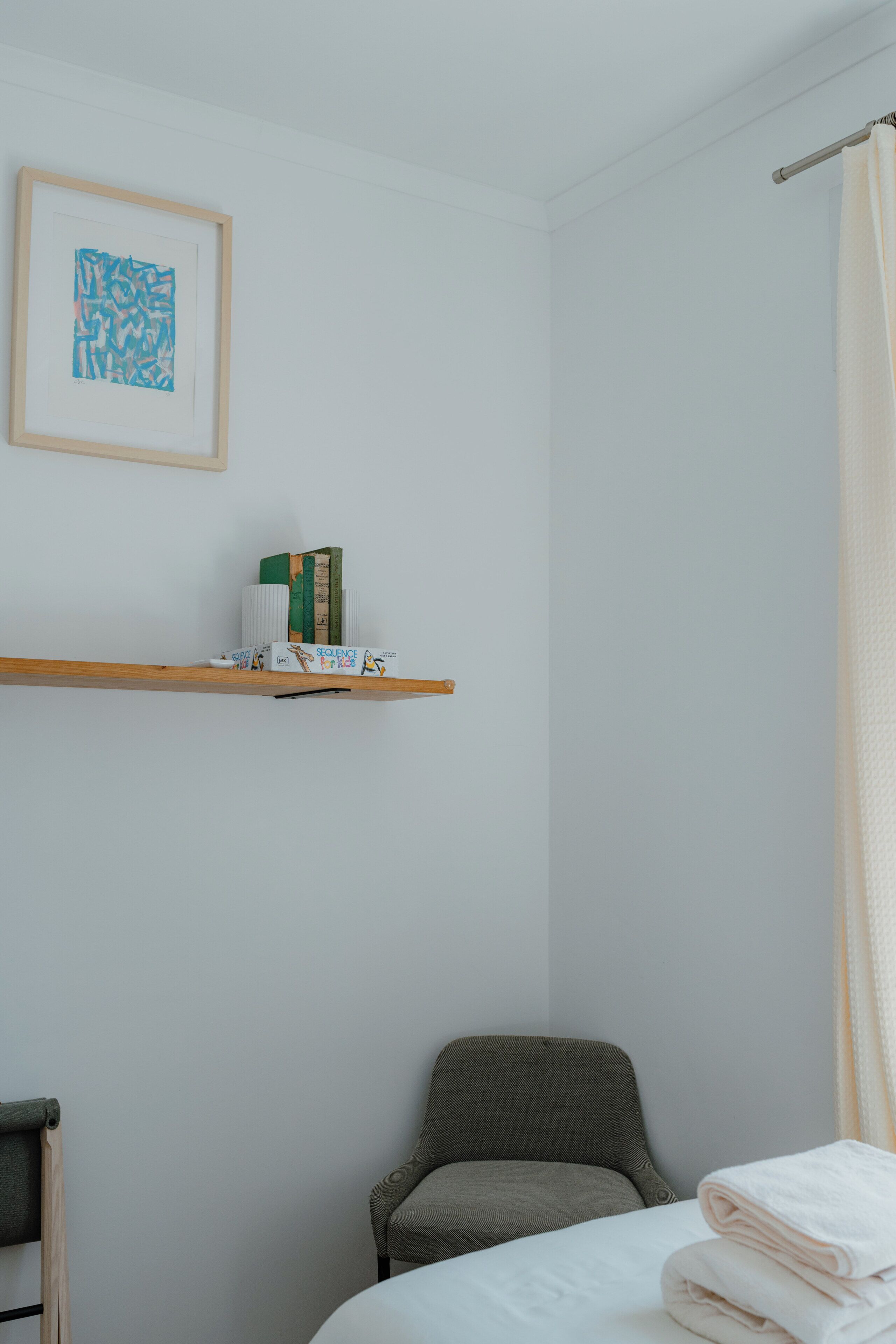 A bedroom with a bed, a chair, a shelf with books, a framed abstract painting, and a shower curtain.
