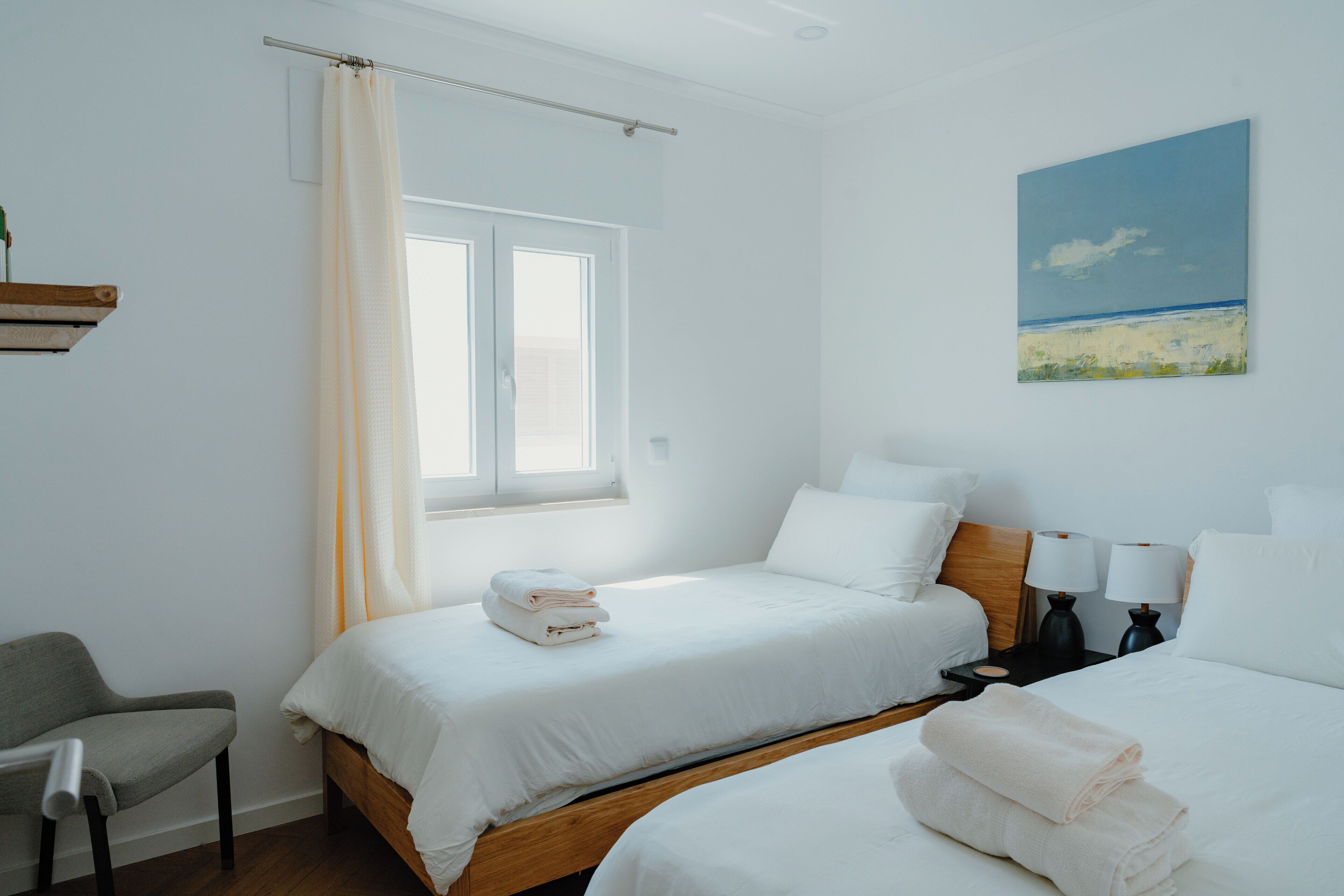 Ein Hotelzimmer mit zwei Betten, einem Sessel, einem Gemälde und einem Fenster mit Vorhängen.