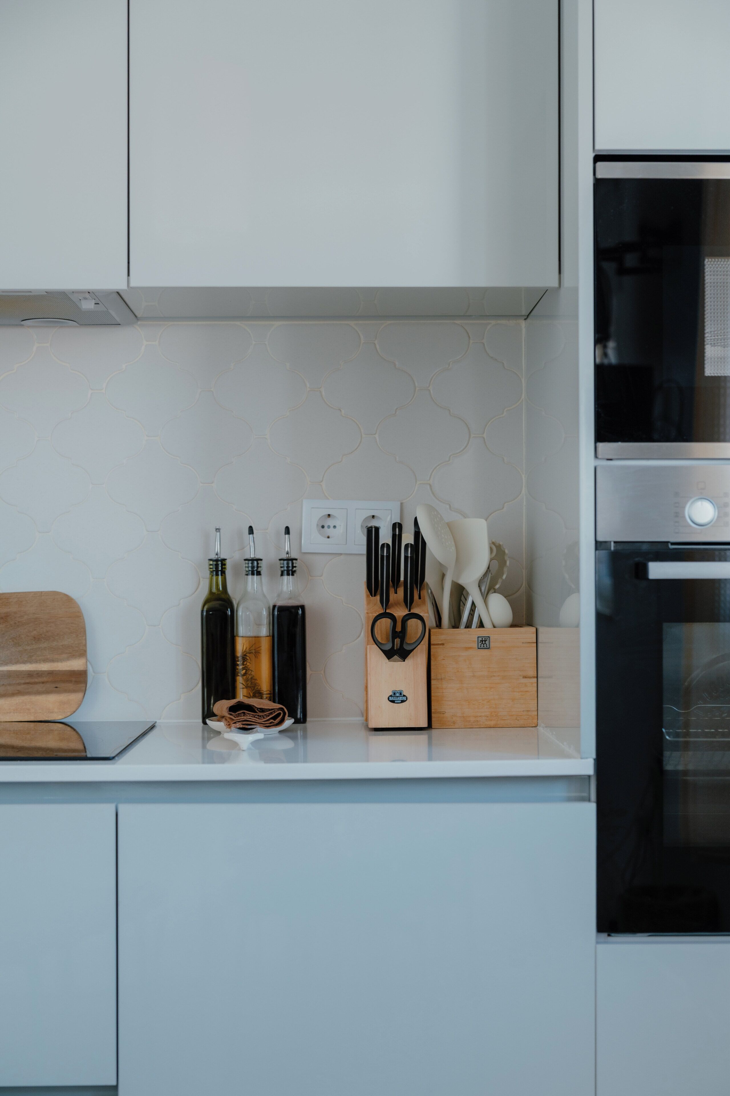 A kitchen with white cabinets, a built-in oven, and a countertop with bottles, a knife block, and a cutting board.