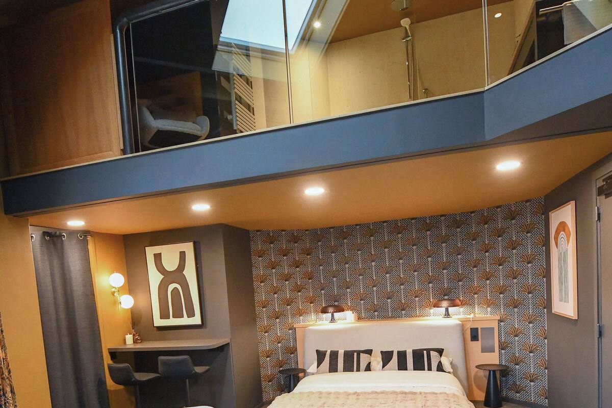 A modern bedroom with a large bed, a seating area with a chair and ottoman, a small table with a glass and candle, and a bathroom with a shower and toilet visible through a glass partition.