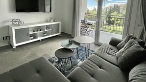 Een moderne woonkamer met een grijze bank, een klein rond tafeltje, een aan de muur bevestigde televisie en een balkon met uitzicht op de omgeving.