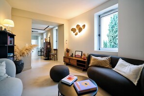 A modern living room with a sofa, a coffee table, a bookshelf, and a dining area in the background. - La Maison des Jardins - Gigondas (Gigondas)