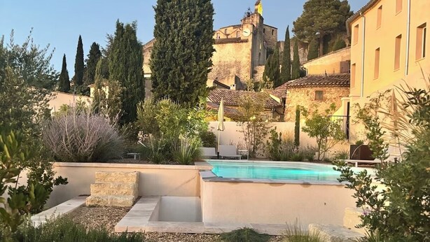 Zona de piscina con solárium de piedra, rodeada de plantas y vistas a un edificio histórico.