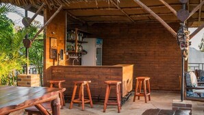 Un espace bar extérieur avec des tabourets en bois et une longue table en bois, entouré de murs en briques et surmonté d’un toit de chaume.