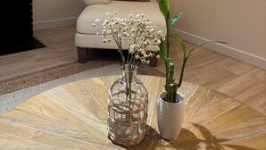 Un salon avec un canapé, un vase en verre rempli de fleurs, une plante en pot, une table en bois, une cheminée, une fenêtre et un tableau accroché au mur.