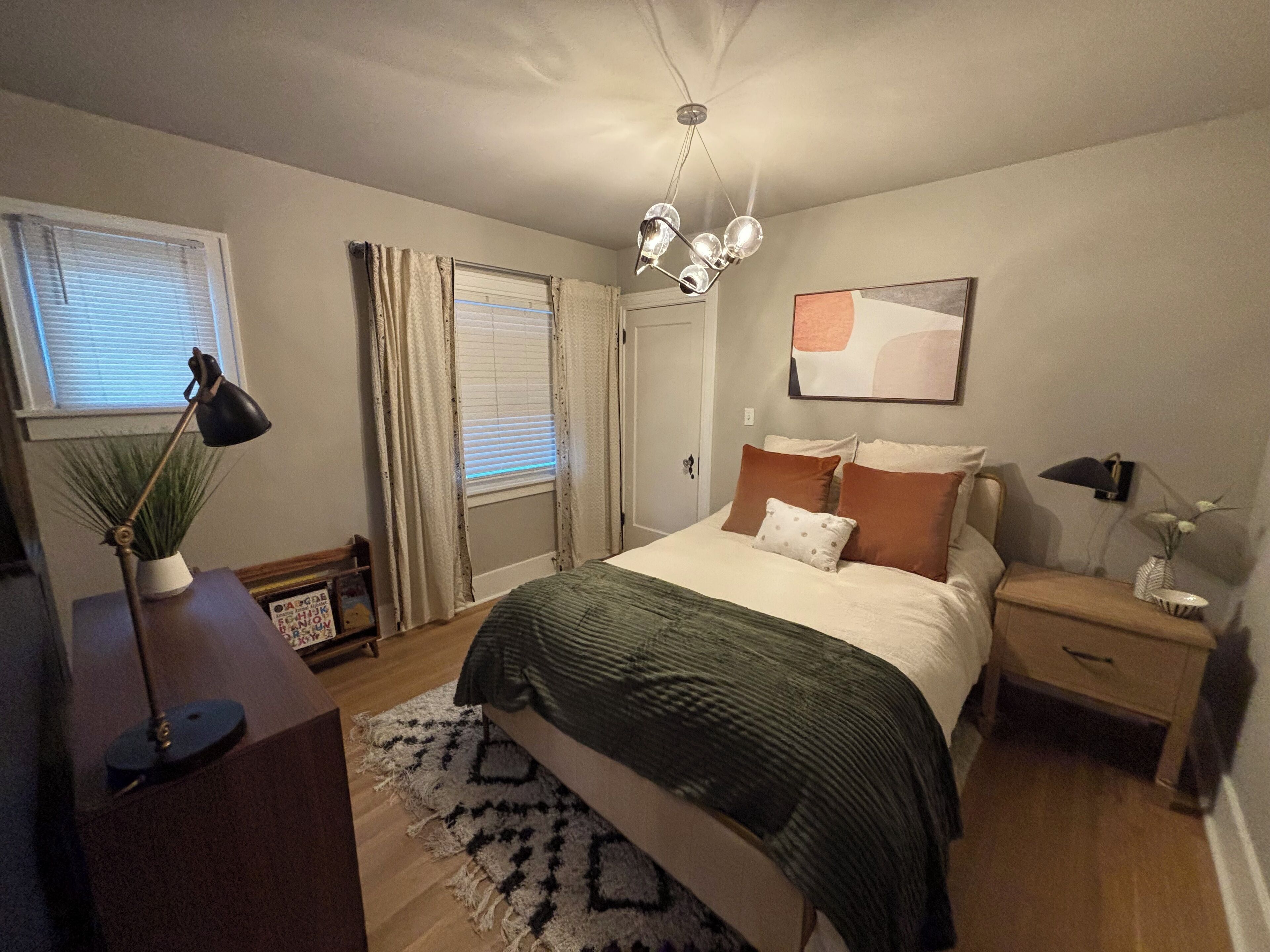 A bedroom with a bed, bedside tables, a desk, a lamp, a plant, and a window with curtains.