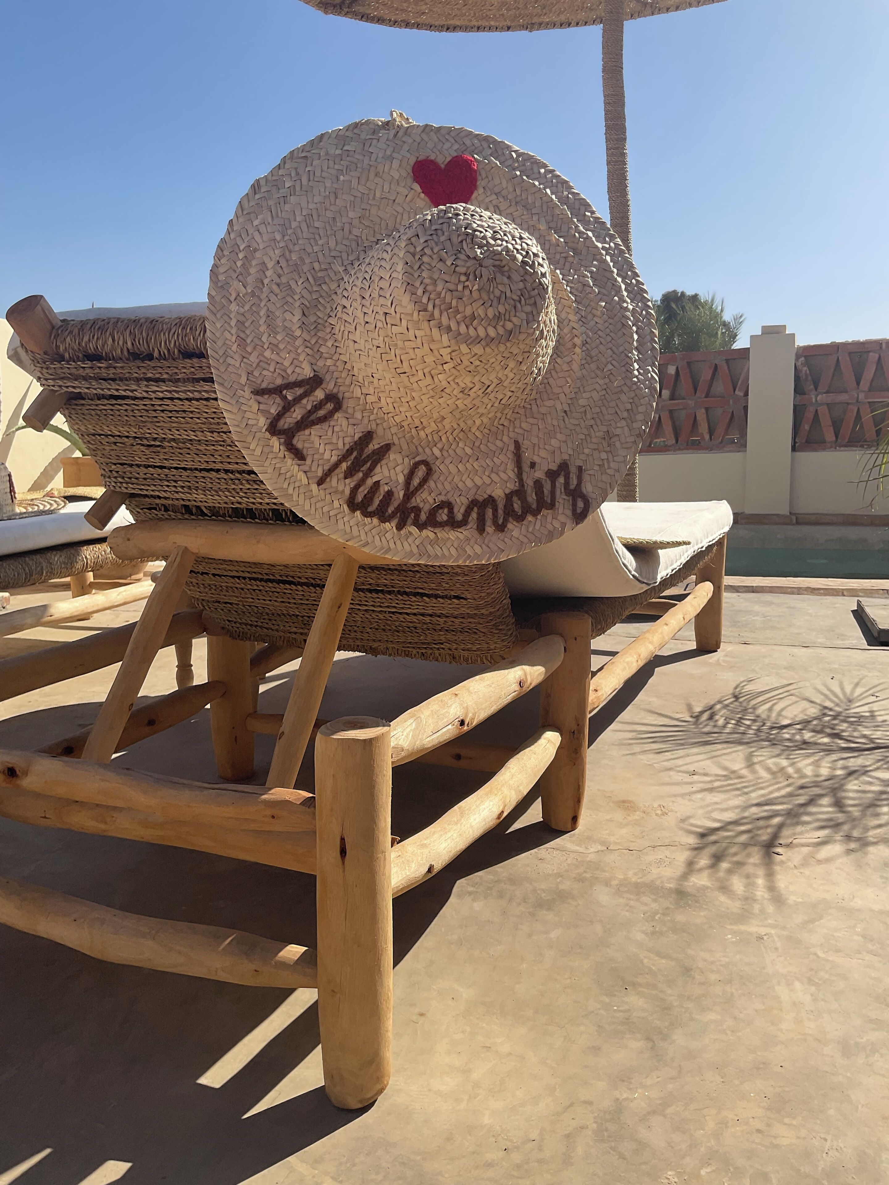 A wooden lounge chair with a woven hat placed on it, and the name "Al Muhandis" written on the hat.
