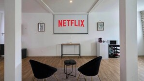 Een moderne woonkamer met een grote tv waarop “NETFLIX” in rode letters staat, twee zwarte stoelen, een klein tafeltje en een witte kast met een magnetron.
