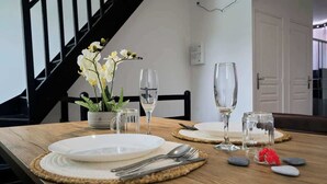 Een eettafel gedekt met witte borden, bestek en glazen, met een vaas witte bloemen en een kleine potplant.