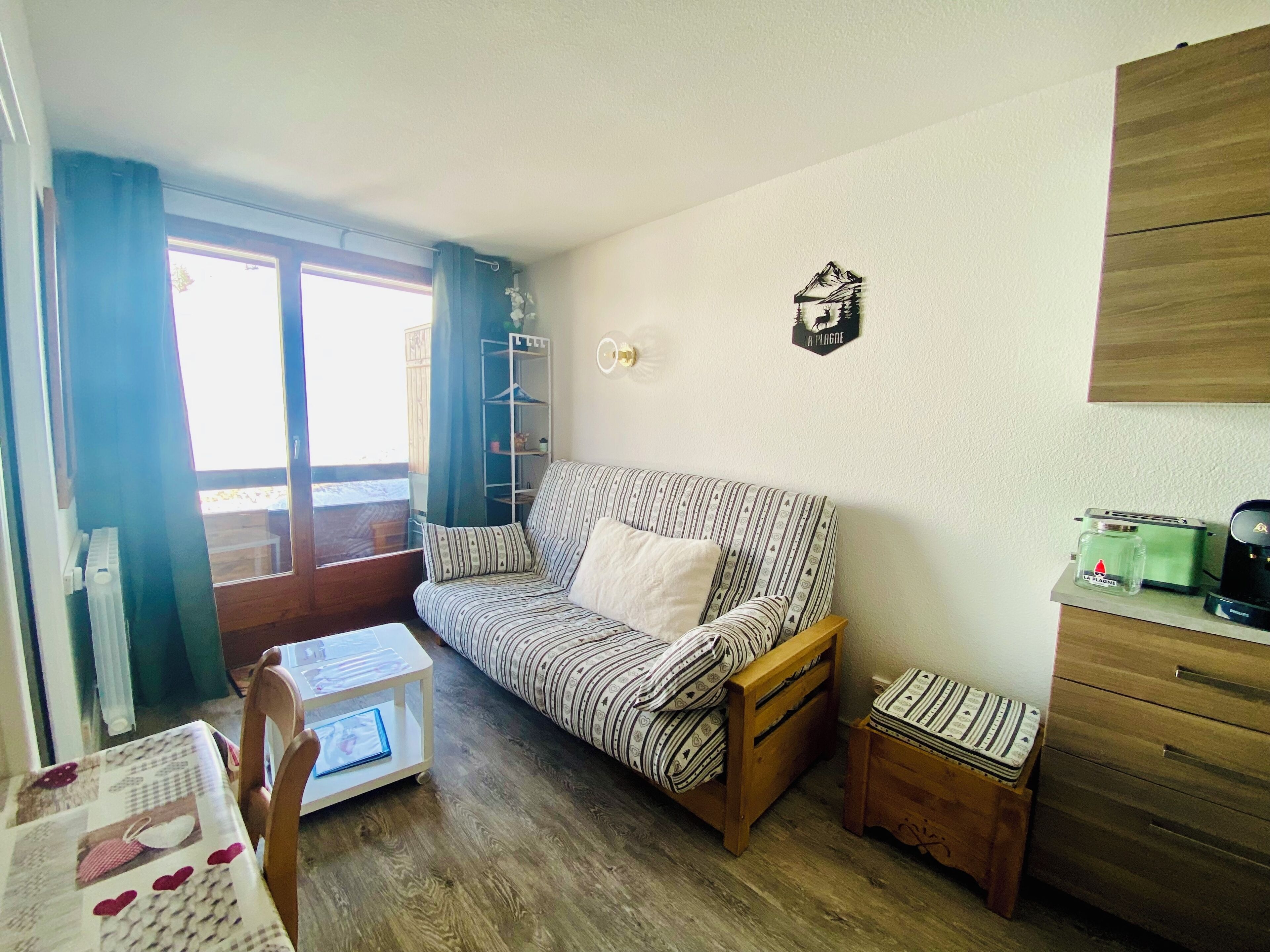 Una habitación acogedora con un sofá a rayas, una pequeña mesa de madera, un radiador, una estantería con objetos y una ventana con cortinas.