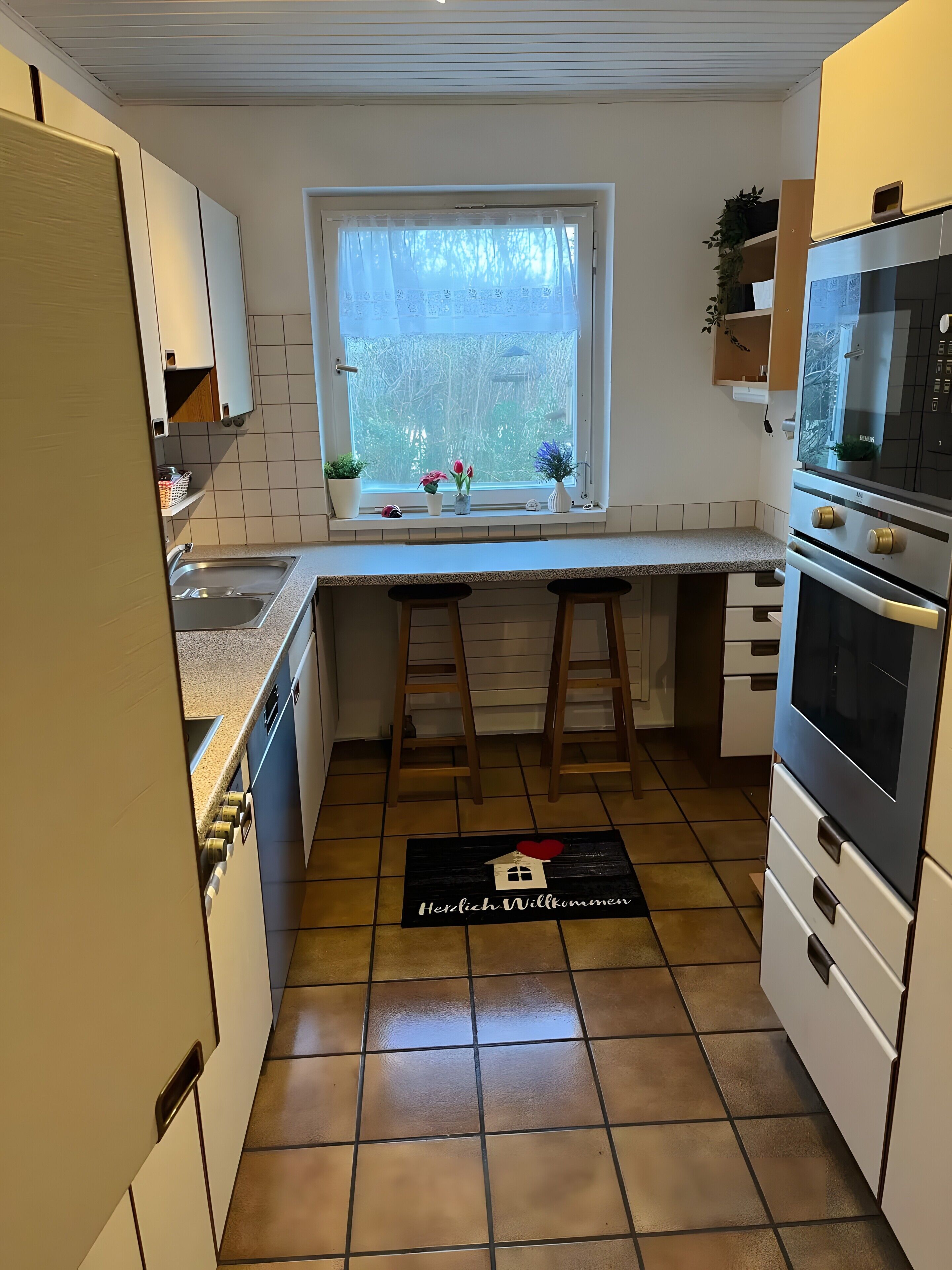 A compact kitchen with a window, a sink, stools, and a welcome mat.