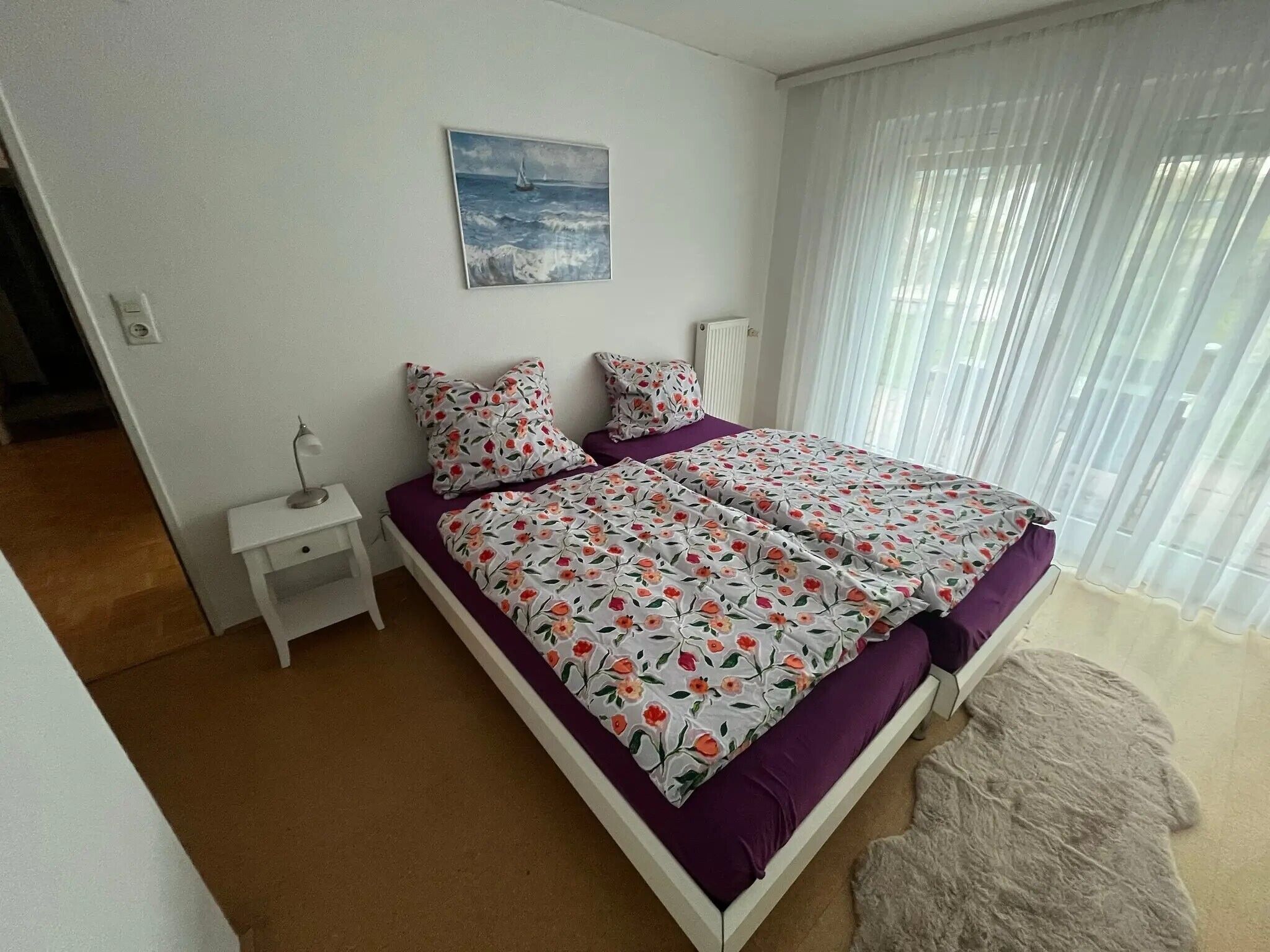 A bedroom with a bed, a nightstand, a painting, and a window with curtains.