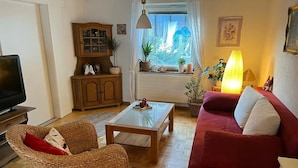Ein Wohnzimmer mit Fernseher, Korbmöbeln, einem roten Sofa und einem Fenster mit Pflanzen.