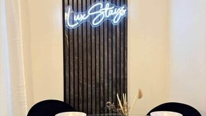 A dining area with a glass table set for four, a vertical wooden panel with a neon sign, and a vase with dried plants. - Modern 4-Bed House • Sleeps 9 •Free Parking, Leeds (Leeds)