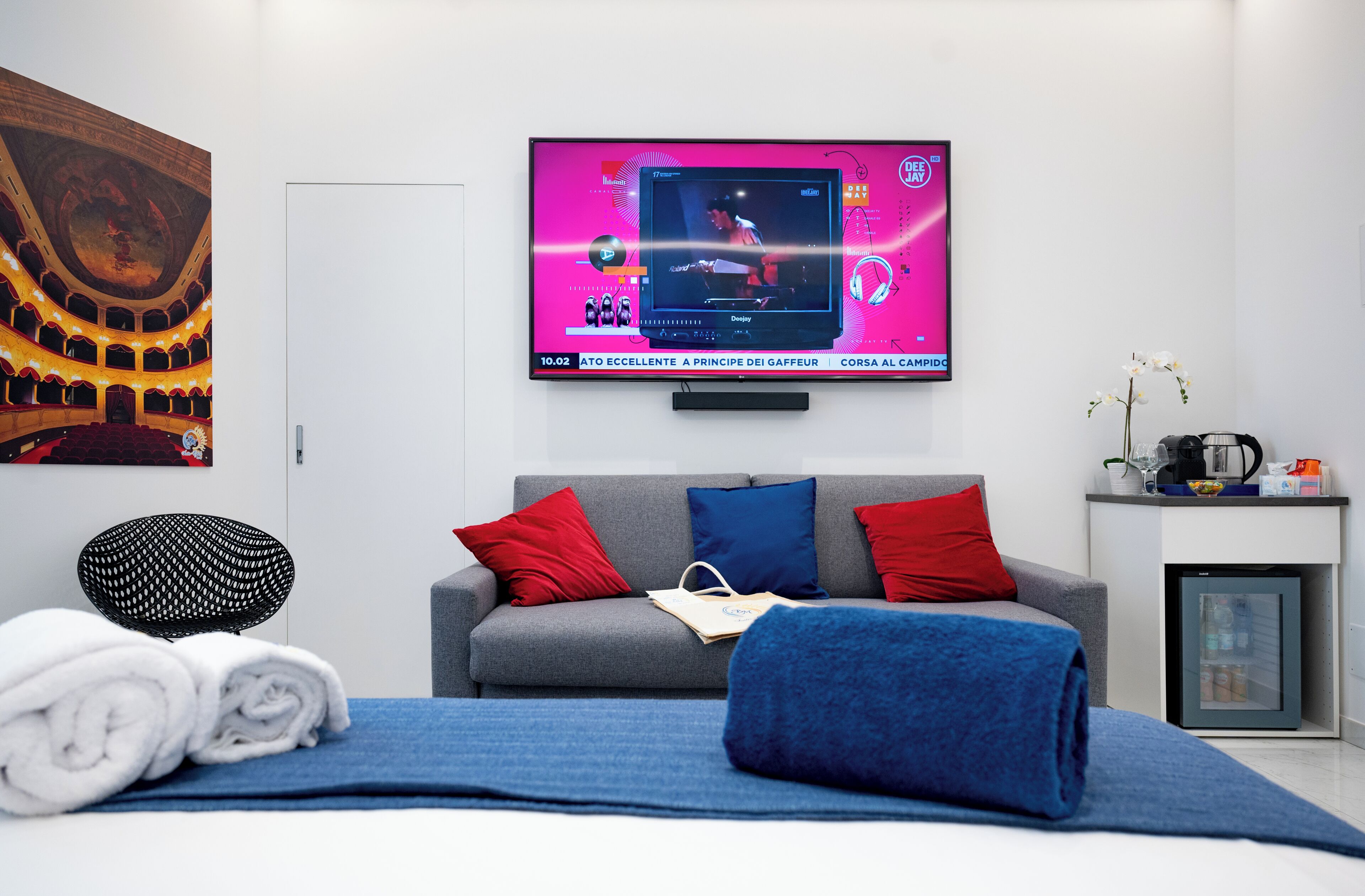 Moderni hotellihuone, jossa on suuri, litteänäyttöinen televisio, jolta näkyy elokuva, harmaa sohva, jossa on punaisia ja sinisiä koristetyynyjä, pieni sivupöytä, jossa on kahvinkeitin, ja seinälle asennettu televisio.