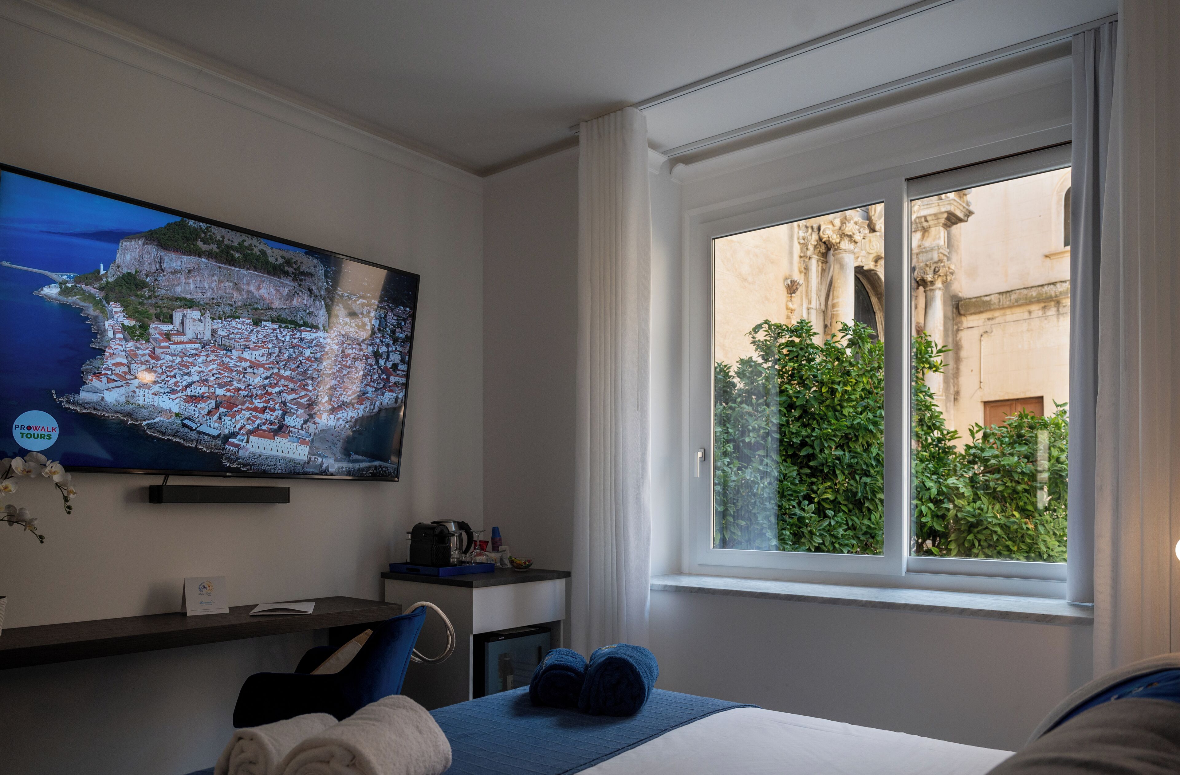 A hotel room with a large TV displaying a scenic view, a bed with blue bedding, a desk with a coffee maker, and a window with a view of greenery and architecture.