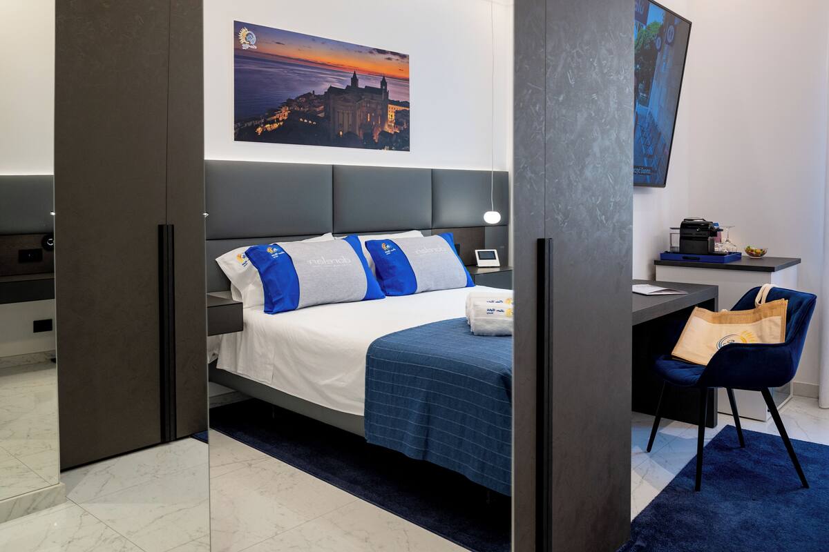 A modern hotel room with a bed, a desk, a chair, and a painting of a sunset over a cityscape.