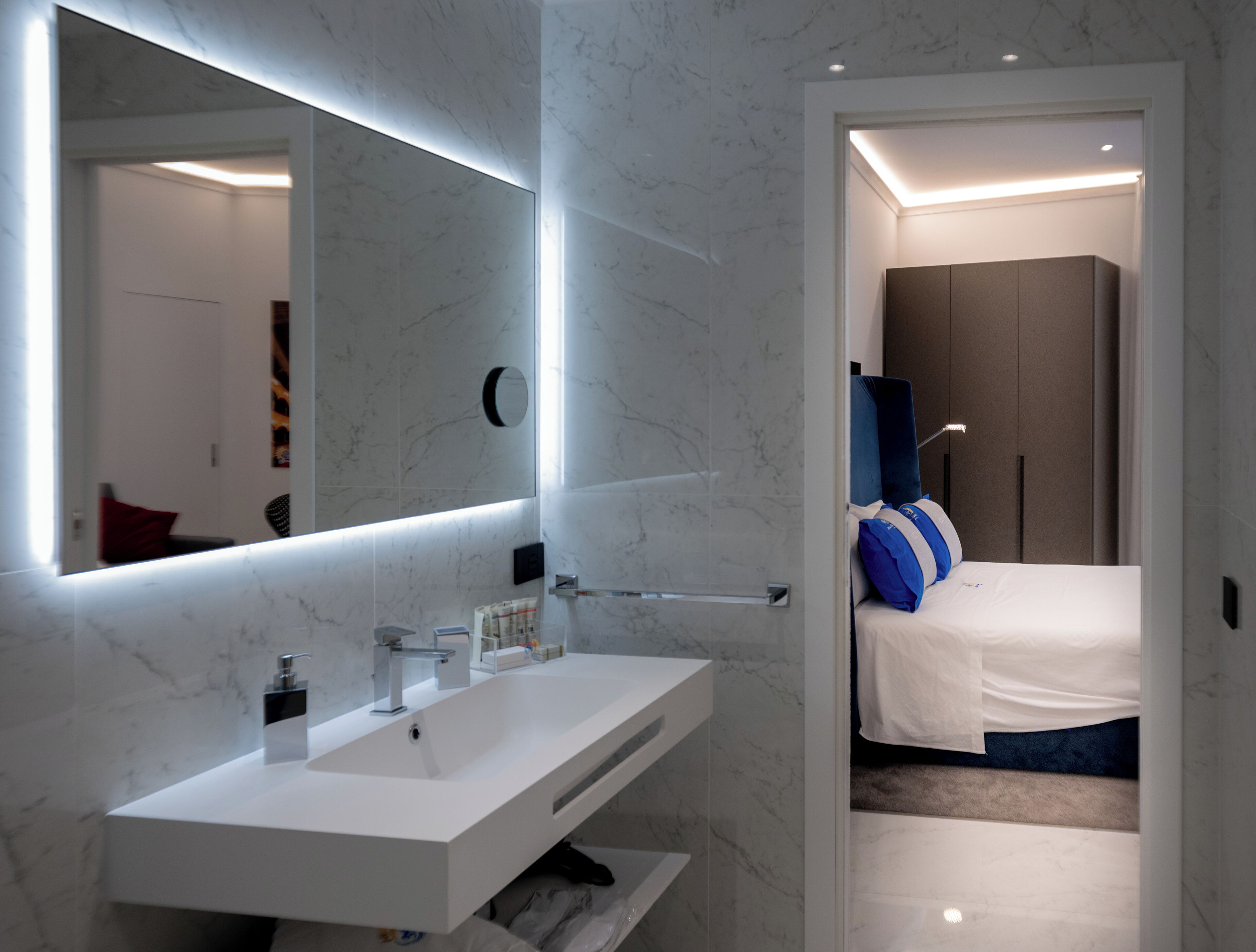 A modern bathroom with a white vanity, a large mirror, and a bed with blue and white bedding in the adjacent room.