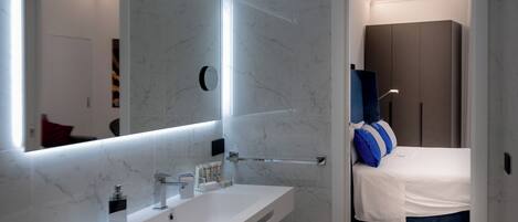 A modern bathroom with a white vanity, a large mirror, and a bed with blue and white bedding in the adjacent room.