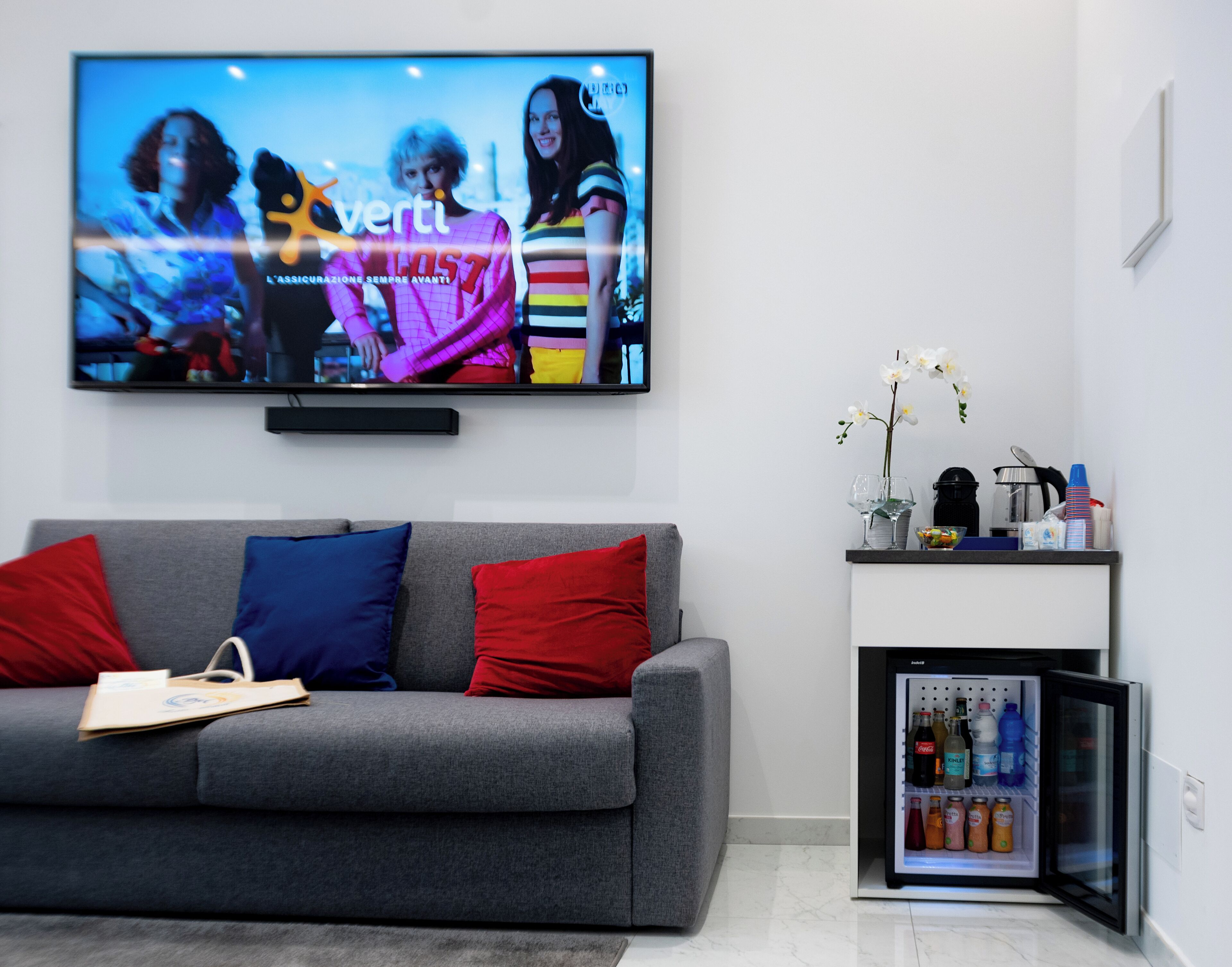 Una sala de estar moderna con un sofá gris, cojines rojos y azules, un televisor de pantalla plana, un mini refrigerador y una pequeña mesa con un jarrón de flores.