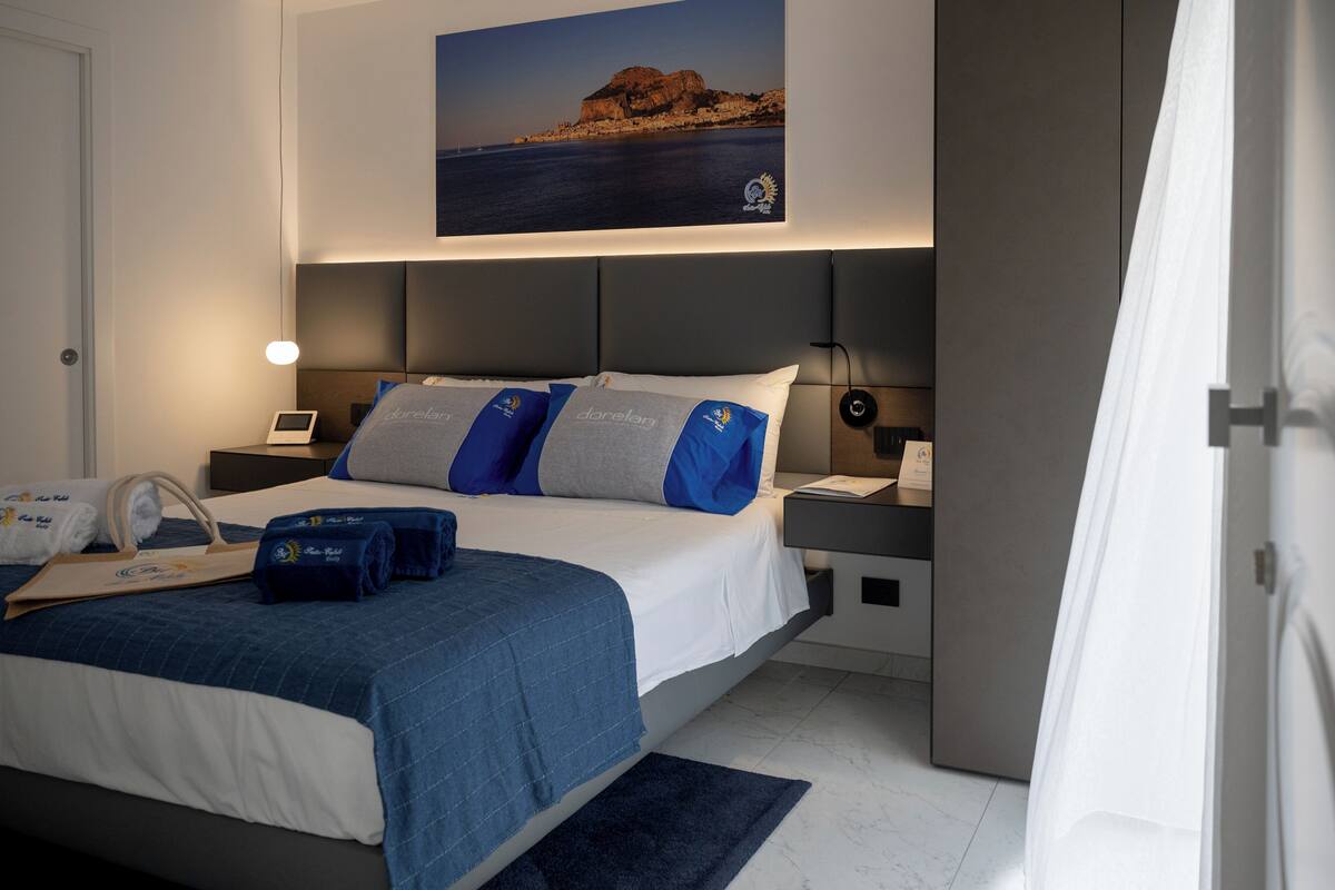 A modern hotel room with a large bed, a nightstand with a lamp, a desk with a chair, and a view of the sea.