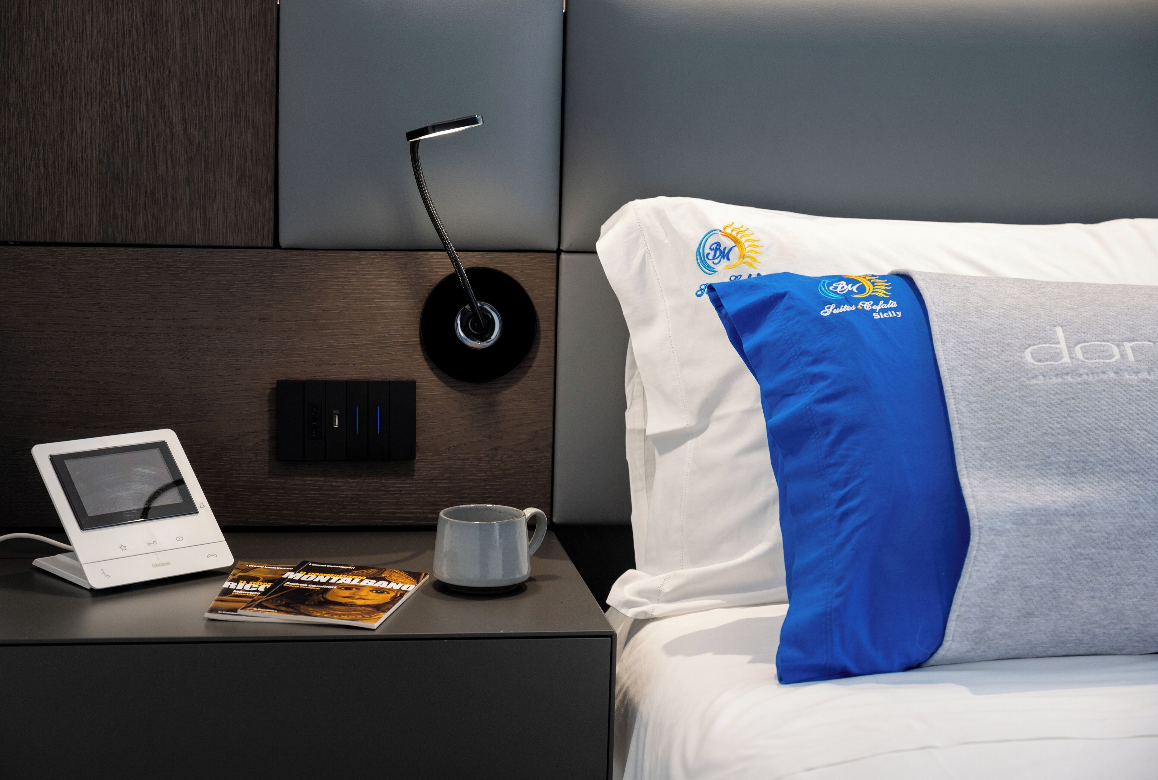 A neatly made hotel room with a bed, a bedside table, a digital clock, a remote control, and a magazine.