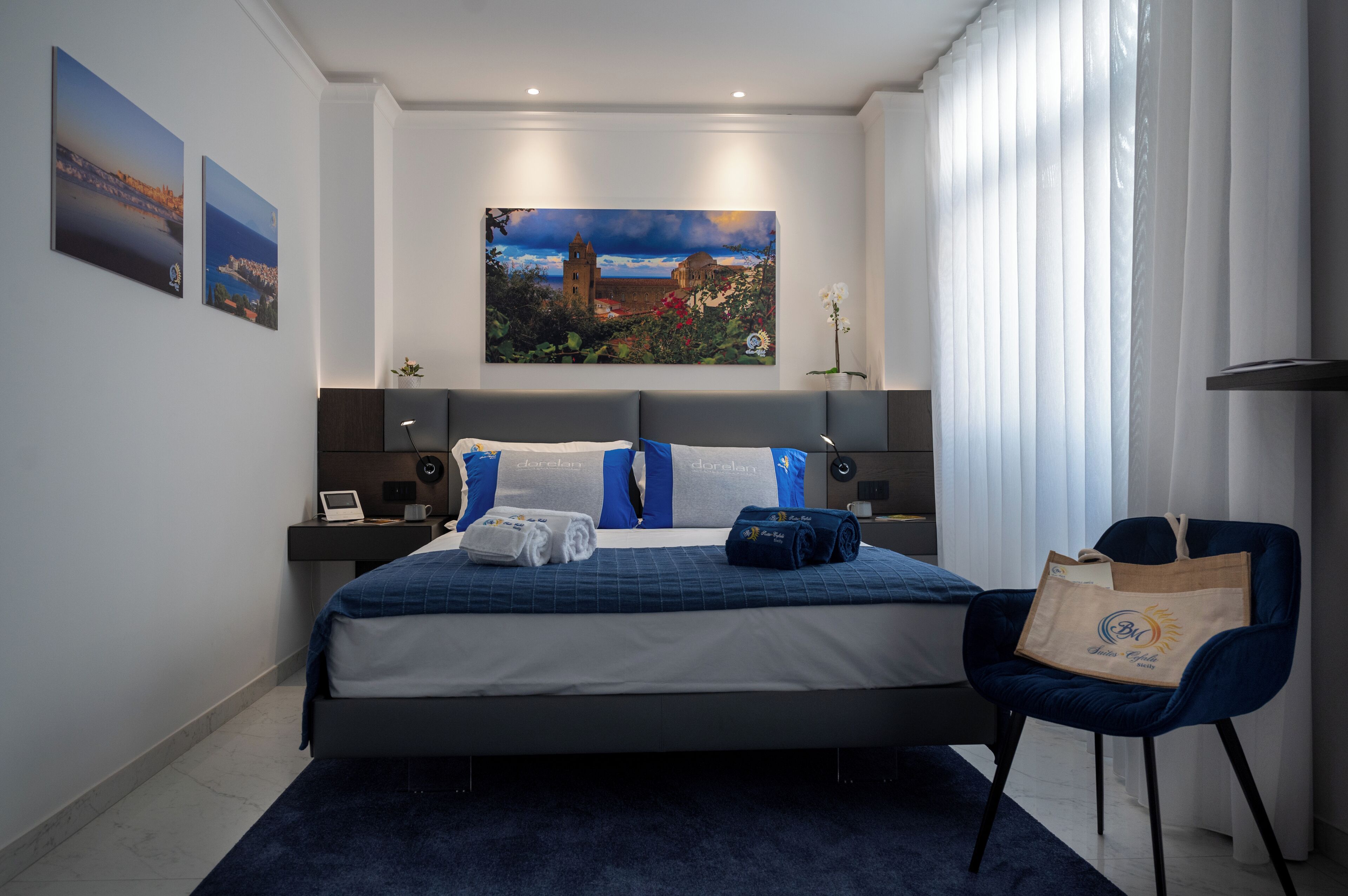 A modern hotel room with a large bed, a desk, and a chair. There are two paintings on the wall, a window with blinds, and a nightstand with a lamp.