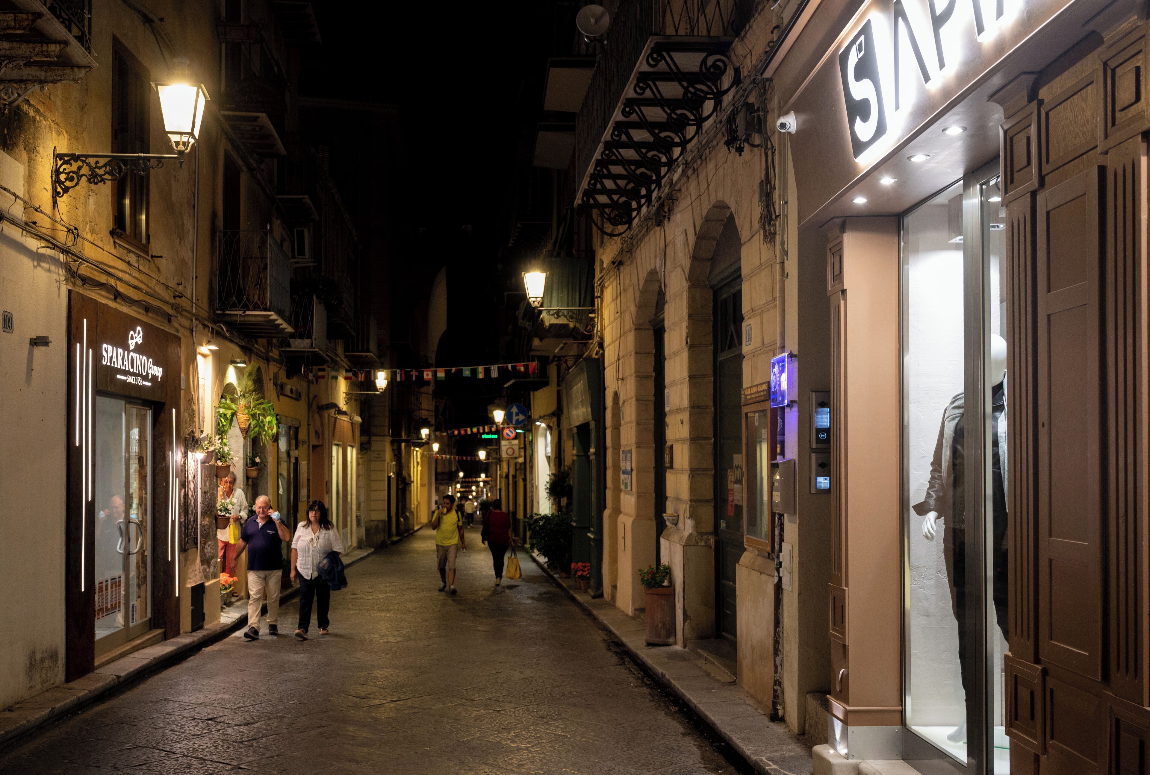 Una calle estrecha bordeada de tiendas y edificios, con una tapa de alcantarilla y un anuncio de un balneario.