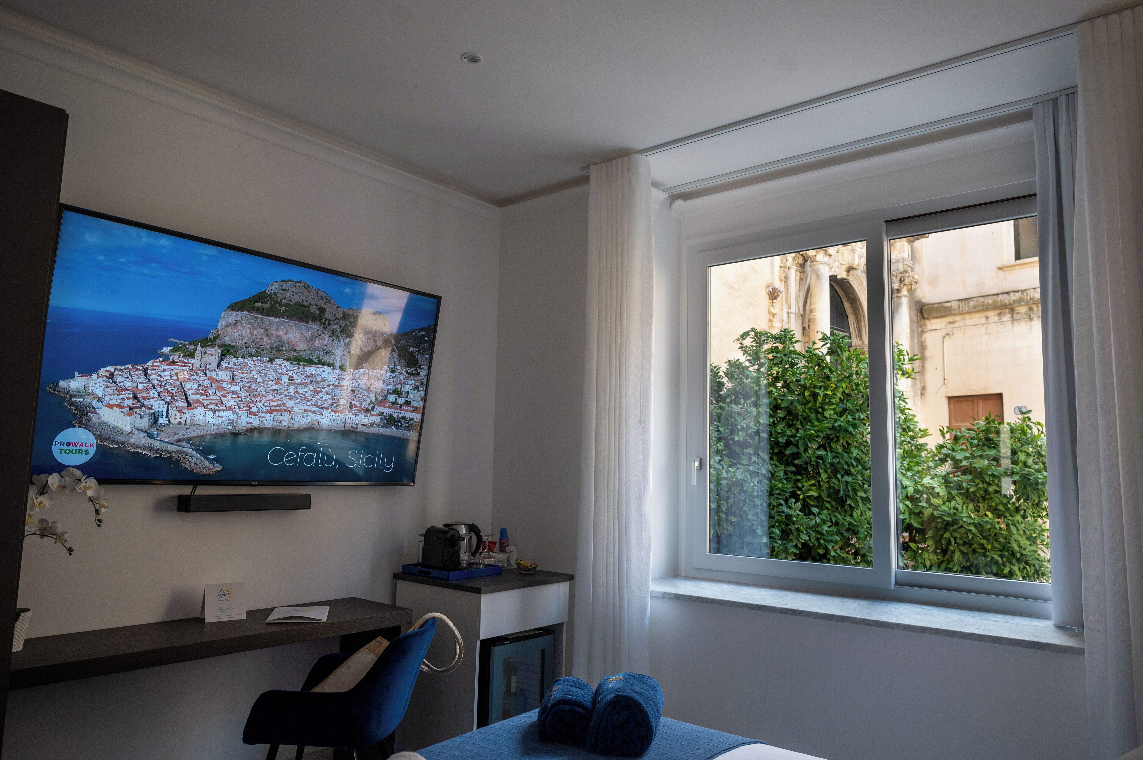 Et hotelværelse med et stort fjernsyn der viser en naturskøn udsigt over byen, et skrivebord med en computer, og et vindue med udsigt til grønt og bygninger.