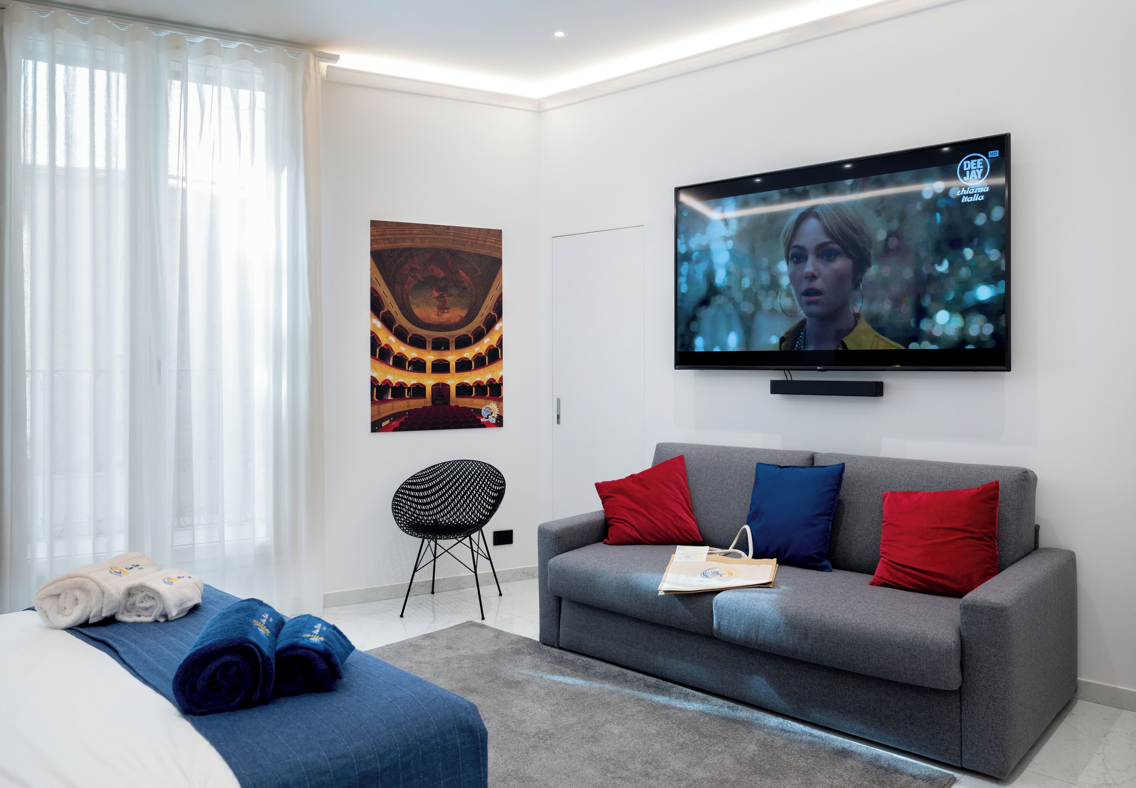 A modern hotel room with a grey sofa, red and blue cushions, a flat-screen TV, a black chair, and a bed with rolled towels.