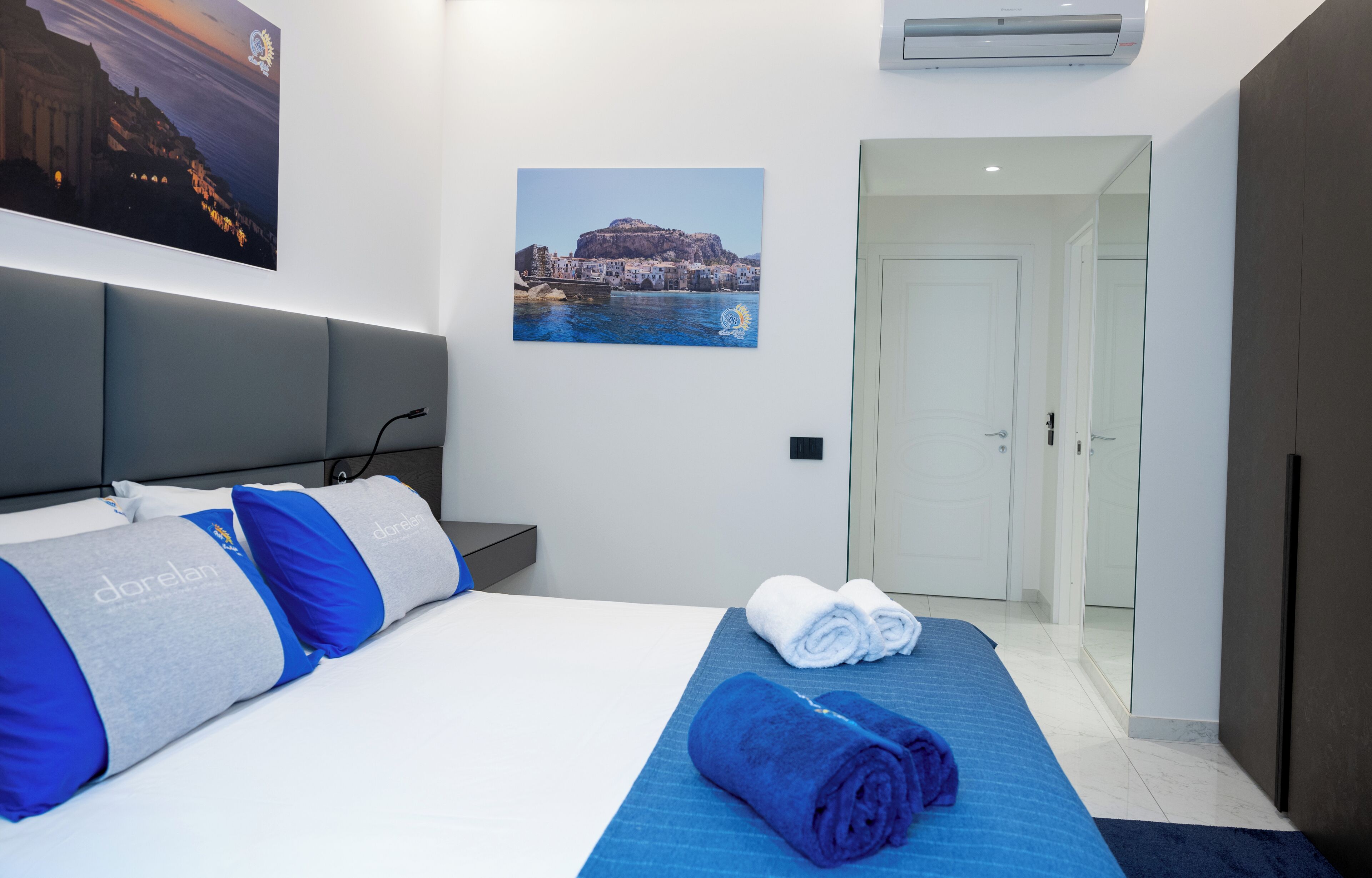 A hotel room with a bed, a blue towel, a rolled-up blue towel, a picture of a coastal town, and a door.