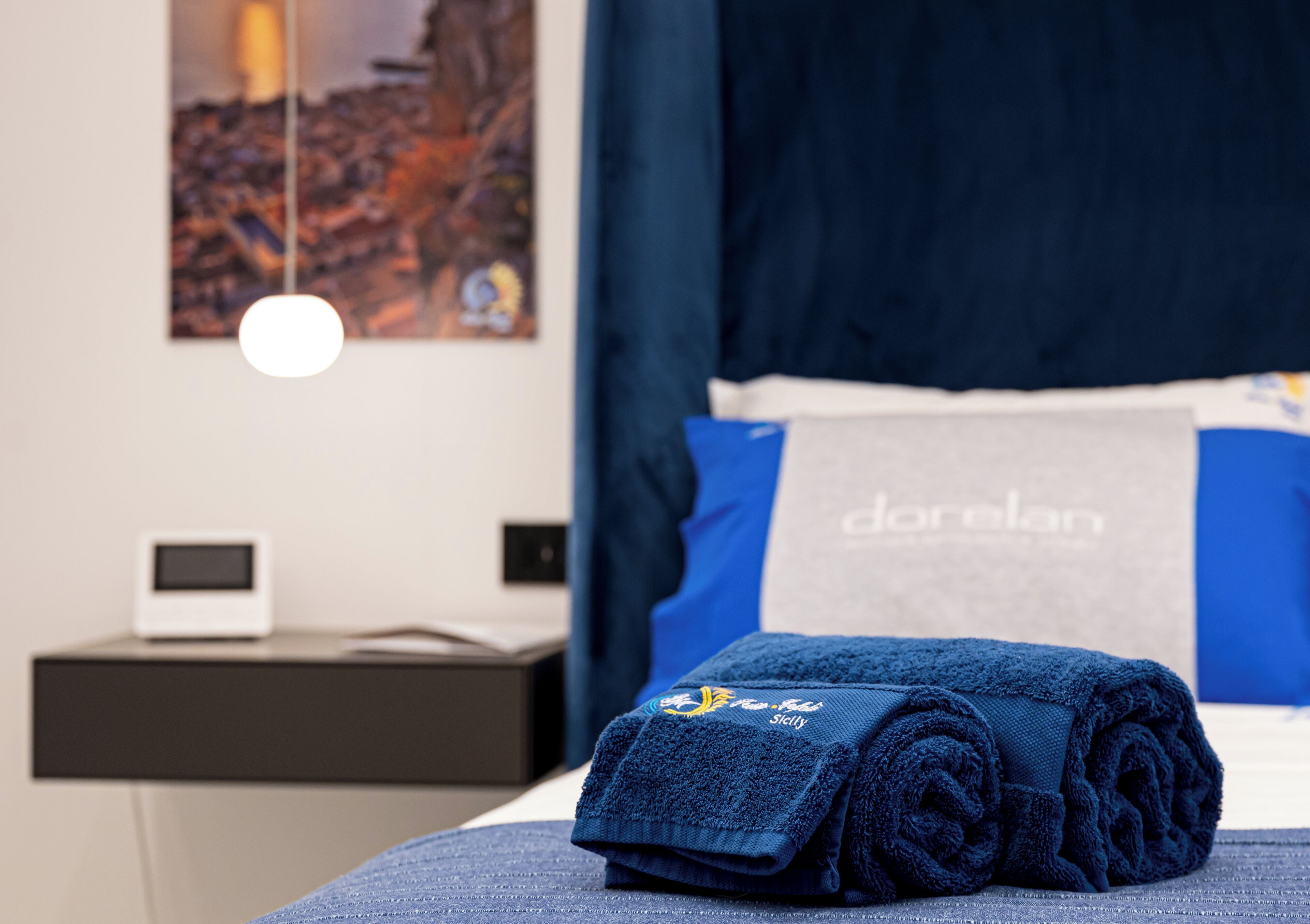 A neatly made bed with a folded towel, a bedside table with a small electronic device, and a wall-mounted light fixture.