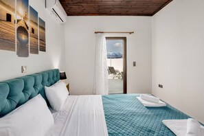 Un dormitorio con cabecera tapizada en color verde azulado, cama con ropa blanca, colcha acolchada azul y vistas a un balcón con mobiliario exterior.