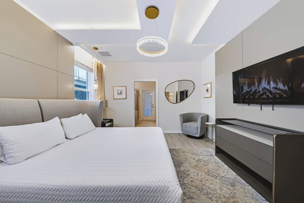 A modern hotel room with a large bed, a flat-screen TV, a seating area with a chair, and a view of the city through the window. - 10BR house with private pool and game room (Clermont)