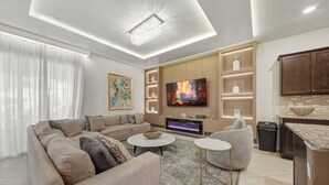 A modern living room with a large sofa, a flat-screen TV, a fireplace, and a kitchen area. - 10BR house at Windsor Cay Resort (Clermont)