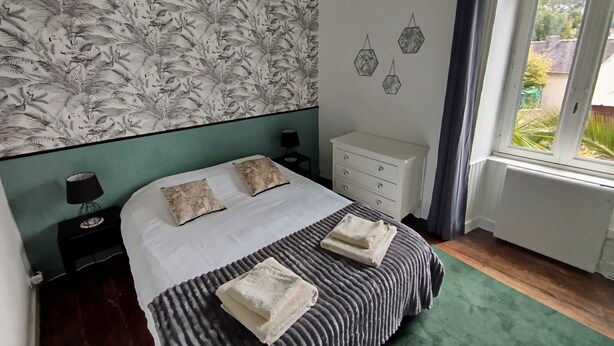 A bedroom with a patterned wallpaper, a bed with two pillows, a bedside table with a lamp, a dresser, and a window with a view of greenery. - Cosy T2 with view in Morlaix between station and town center (Morlaix)