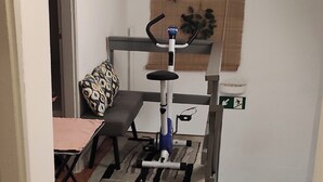 Een compacte kamer met een klein hometrainer, een bank, een eettafel en een raam met een zonwering.