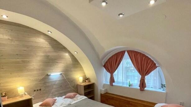 Een slaapkamer met een gebogen plafond, een houten hoofdbord, een groot raam met gordijnen en een nachtkastje met een lamp.