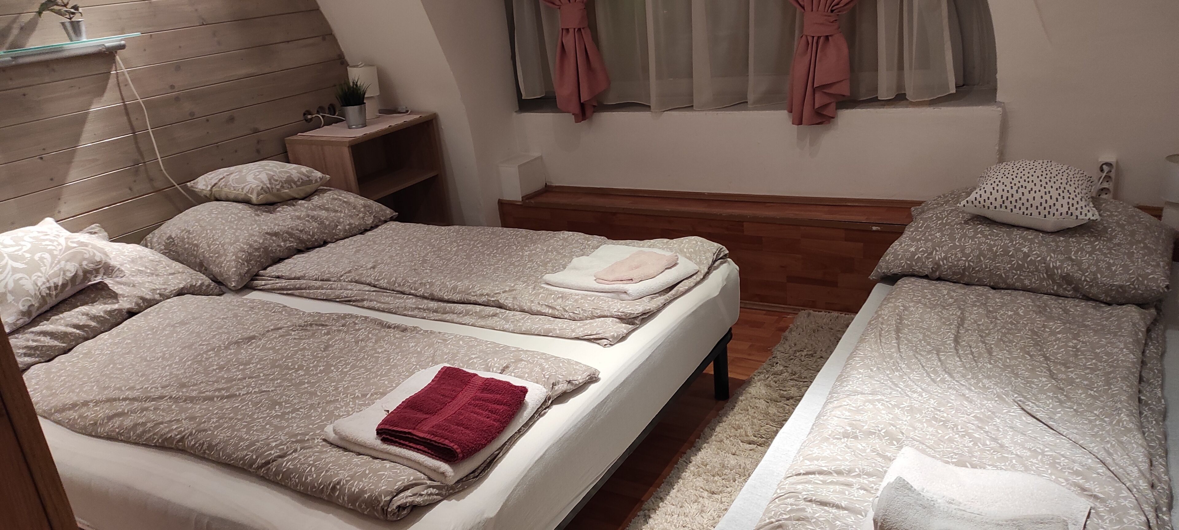 Un dormitorio con dos camas, una mesita de noche, una ventana con cortinas y un cabecero de madera.