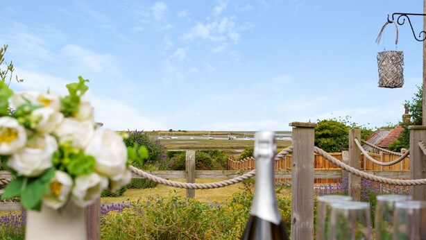A garden setting with a wooden fence, a rope barrier, and a bottle of wine with glasses. - Sunnydene - sleeps 6 guests  in 3 bedrooms (Brancaster Staithe)