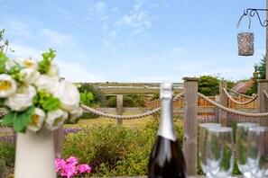A garden setting with a wooden fence, a rope barrier, and a bottle of wine with glasses. - Sunnydene - sleeps 6 guests  in 3 bedrooms (Brancaster Staithe)