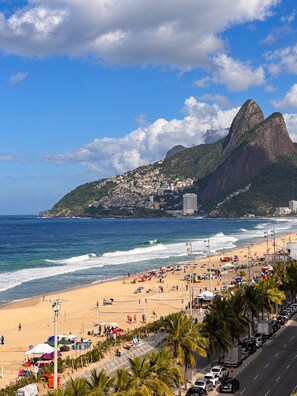 A beach with a mountain in the background, a road with parked cars, and umbrellas on the sand. - Poolfront with Ocean View (Rio de Janeiro)