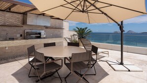 Patio esterno con tavolo e sedie rotonde, un ombrellone grande, un barbecue e vista sul mare.