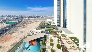 A high-rise building with a modern design, a landscaped area with palm trees, and a road with vehicles. - Modern 2BR Haven in Golf Gate Pool & Gym Access (Dubai)