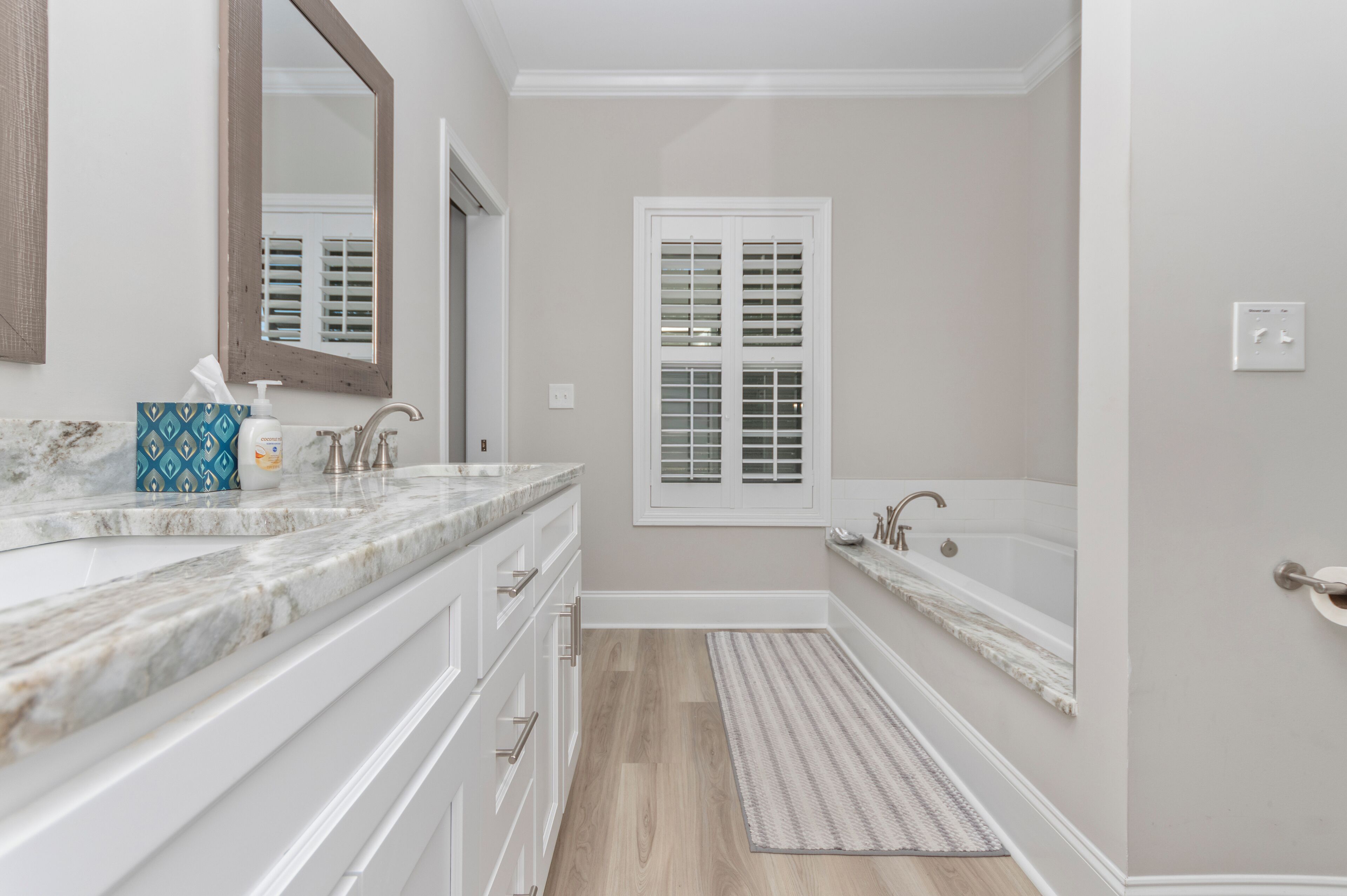 Une salle de bain avec un comptoir en marbre, un grand miroir, une fenêtre avec des stores, une baignoire et un plancher en bois.