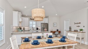 A modern kitchen with white cabinetry, a central island, and a dining area set for six with blue tableware. - Coastal Comfort ~ your own Heated Pool ~ Golf Cart (North Myrtle Beach)