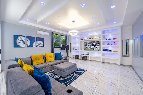 Una sala de estar moderna con un sofá seccional gris, cojines amarillos y azules, un televisor grande y un estante empotrado con diversos objetos.