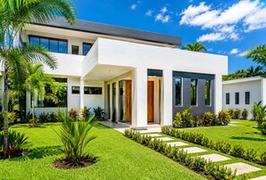 Una casa moderna de dos pisos con un porche cubierto, un césped cuidado y diversas plantas.