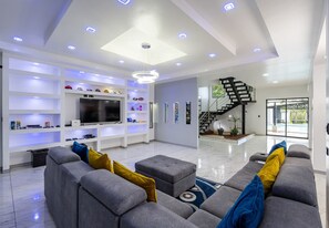 A modern living room with a grey sofa, a flat-screen TV, and shelves with various items. - Villa Montreal | Private Pool + Game Room (La Fortuna)