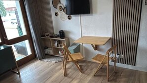 Una stanza con pavimento in legno, un piccolo tavolo in legno, due sedie in legno, una TV montata a parete e una finestra con tende.